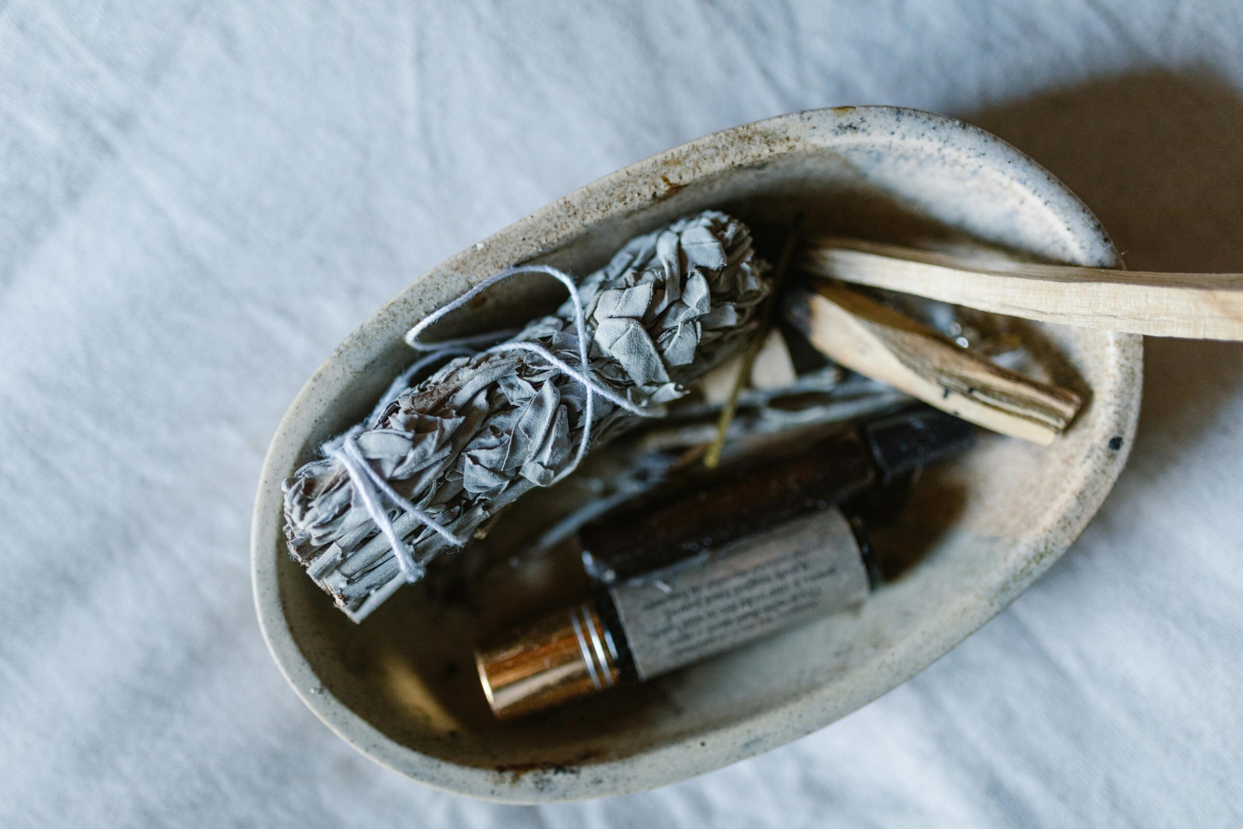 A serene still life featuring a bundle of sage, essential oils, and wooden sticks in a rustic bowl.